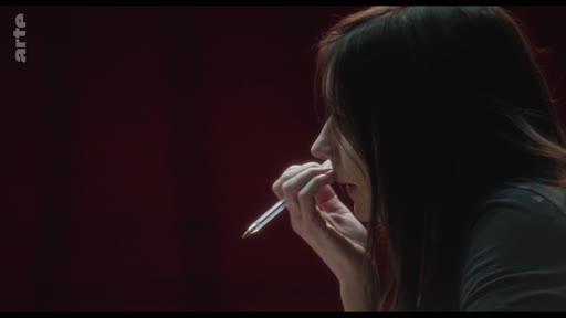 Une femme en gros plan, vue de profil, semble plongée dans une réflexion intense tout en tenant un stylo près de ses lèvres, illustrant une séquence sur les attentes sociétales.