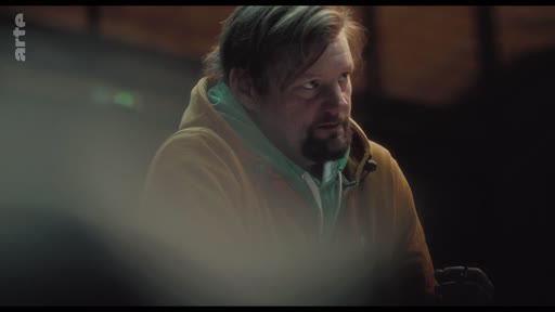 Un homme avec une barbe et une coiffure décontractée, vêtu d'un sweat-shirt à capuche, est filmé en gros plan dans une ambiance tamisée. Il semble écouter ou observer une discussion, reflétant une atmosphère intime ou réflexive.