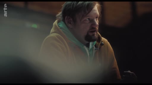 Un homme barbu portant un sweat-shirt à capuche s'exprime face caméra lors d'une interview. Il semble discuter de sujets de société dans un cadre sombre.