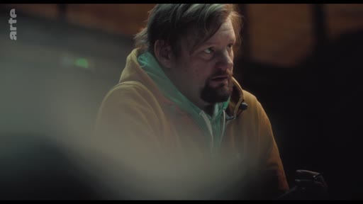 Un homme d'âge moyen avec une barbe et un sweat-shirt à capuche est filmé en plan rapproché lors d'une discussion, probablement dans le cadre d'une interview ou d'un documentaire.