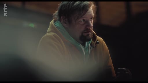 Un homme d'âge moyen avec une barbe et un sweat-shirt à capuche est filmé en gros plan dans une ambiance tamisée. Il semble écouter ou participer à une discussion, son regard étant tourné vers la droite du cadre.