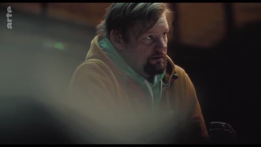 Un homme d'âge moyen avec une barbe et un sweat-shirt à capuche est filmé en plan rapproché lors d'une discussion, illustrant une séquence sur les attentes sociétales.