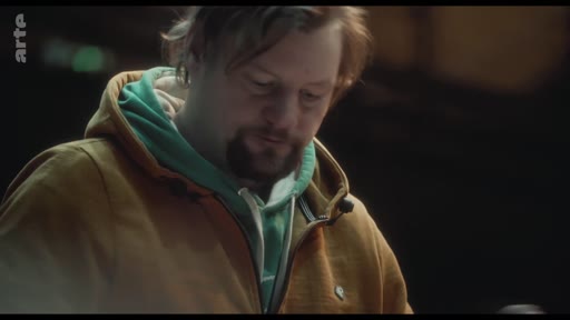 Un homme barbu, vêtu d'une veste jaune et d'un sweat à capuche vert, est filmé en gros plan dans une séquence illustrant la vulnérabilité.