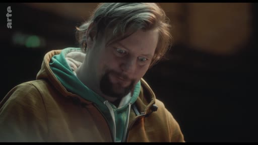 Un homme portant un sweat-shirt à capuche jaune et vert affiche une expression de vulnérabilité et de réflexion, capturé dans un plan rapproché.