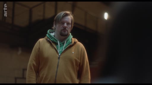 Un homme d'âge moyen, vêtu d'un sweat-shirt jaune à capuche verte, est filmé en plan moyen dans un intérieur sombre, probablement lors d'une discussion sur la situation de son fils.