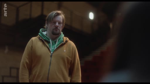 Un homme d'âge moyen, vêtu d'un sweat-shirt à capuche jaune sur un sweat vert, est filmé en plan moyen dans un intérieur sombre, probablement lors d'une scène de dialogue dramatique.