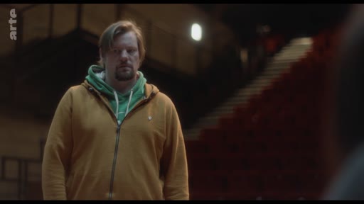 Un homme portant un sweat-shirt à capuche jaune et vert se tient debout dans une salle de théâtre ou de spectacle plongée dans la pénombre, lors d'une discussion sur l'impact sur leur fils.
