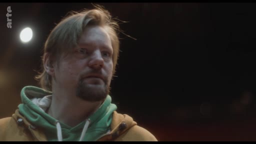 Un homme barbu portant un sweat-shirt à capuche vert et une veste jaune, filmé en gros plan, semble écouter ou observer quelqu'un lors d'une conversation.