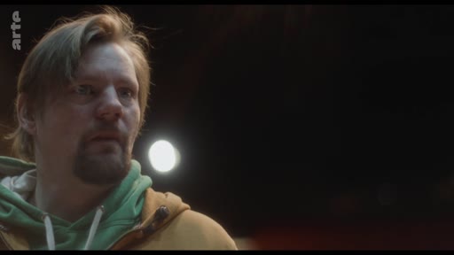 Un homme barbu portant un sweat-shirt à capuche vert et une veste jaune exprime de l'inquiétude ou de la réflexion lors d'une discussion.