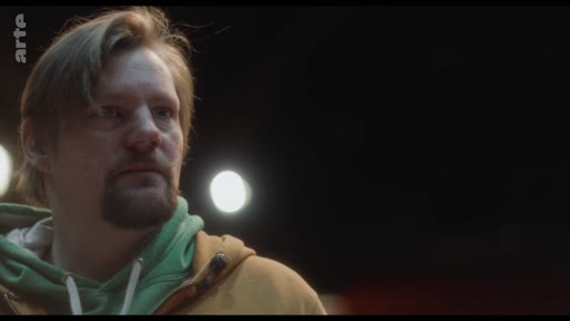 Un homme barbu portant un sweat-shirt à capuche jaune et vert, filmé en gros plan, semble écouter ou observer quelqu'un lors d'une discussion sur les relations de couple.