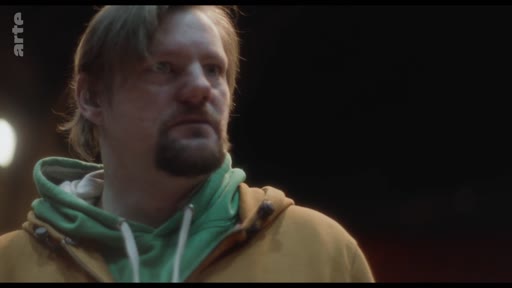 Un homme portant un sweat-shirt à capuche jaune et vert exprime une inquiétude ou une réflexion lors d'une discussion.