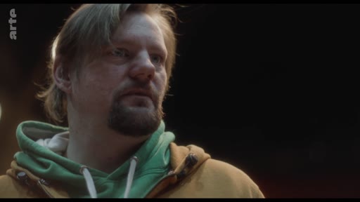 Un homme d'âge moyen avec une barbe et des cheveux mi-longs, vêtu d'un sweat-shirt à capuche vert et jaune, est filmé en gros plan lors d'une discussion.