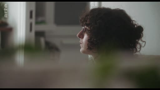 Une femme aux cheveux bouclés, vue de profil, est en train de discuter dans un intérieur lumineux.