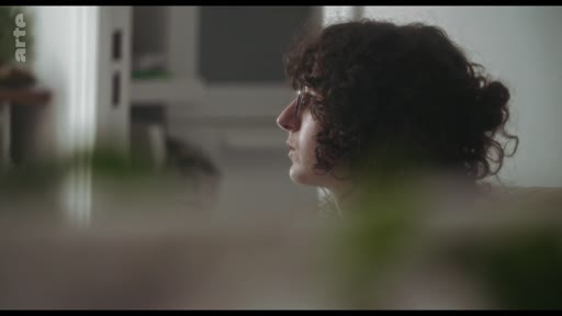 Une femme aux cheveux bruns bouclés est filmée de profil dans un intérieur, lors d'une discussion sur un roman.