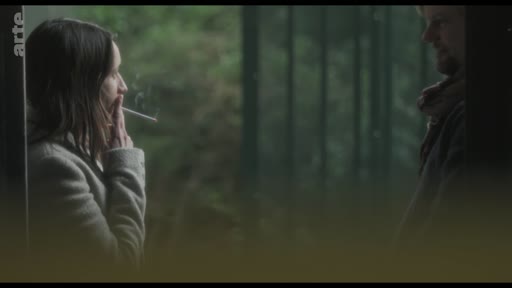 Une femme fume une cigarette tout en regardant un homme dans une scène de dialogue en extérieur, évoquant une atmosphère mélancolique ou dramatique.