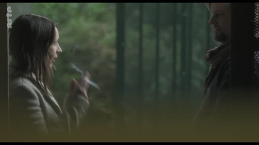 Une femme et un homme se font face lors d'une discussion, la femme tient une cigarette à la main. La scène se déroule dans un cadre sombre et intimiste, évoquant une interaction personnelle ou dramatique.