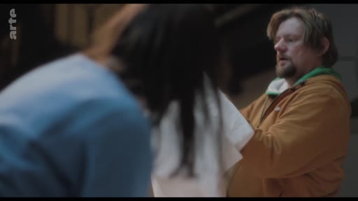 Un homme barbu portant un sweat-shirt orange interagit avec une femme de dos dans ce qui semble être un studio de répétition ou un espace de travail artistique.