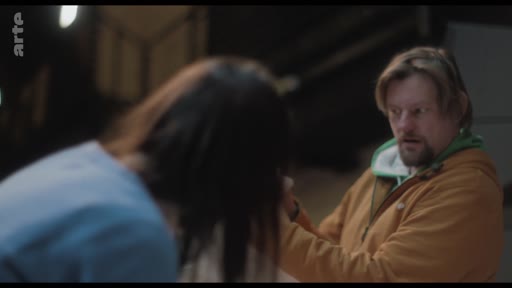 Un homme barbu portant un sweat-shirt à capuche orange discute avec une femme de dos dans ce qui semble être un studio de répétition ou un espace de travail artistique.