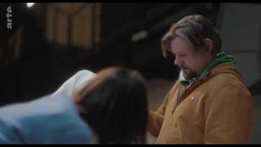 Un homme barbu portant une veste moutarde et un sweat à capuche vert est en pleine discussion avec une personne vue de dos dans ce qui semble être un espace de répétition théâtrale ou un studio.