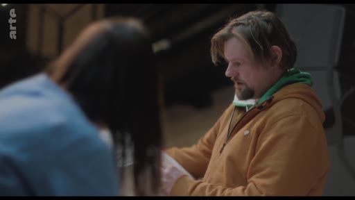 Un homme barbu portant un sweat-shirt à capuche orange discute avec une personne de dos dans ce qui semble être une salle de répétition. La scène montre une interaction de travail ou de préparation artistique.