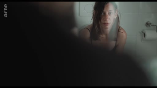 Une femme se regarde dans un miroir de salle de bain, dans une scène illustrant des réflexions sur une relation amoureuse. L'image capture un moment d'introspection personnelle dans un cadre domestique.