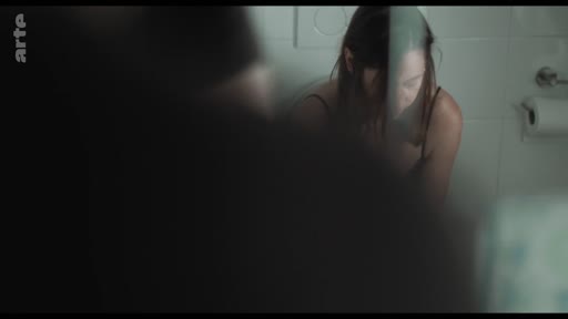 Une femme est assise dans une salle de bain, son visage étant partiellement visible à travers un reflet sur une surface vitrée ou un miroir, évoquant une atmosphère d'intimité ou de réflexion personnelle.