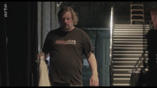 Un homme, possiblement un technicien ou un artiste, se déplace dans les coulisses sombres d'une salle de spectacle, tenant un vêtement à la main. La scène illustre les préparatifs techniques ou artistiques en coulisses.