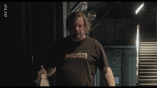 Un homme aux cheveux mi-longs, vêtu d'un t-shirt marron, est filmé dans un environnement sombre rappelant les coulisses d'un théâtre ou d'un plateau de tournage lors des préparatifs.