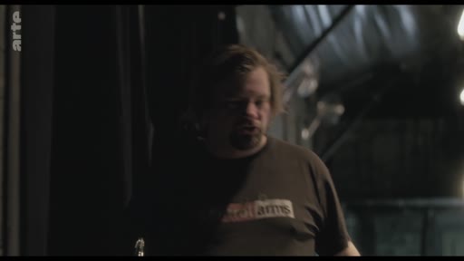 Un homme barbu, possiblement un artiste ou un technicien, est filmé dans un environnement sombre lors de préparatifs de scène. L'éclairage tamisé met en avant son visage et son t-shirt portant une inscription partiellement lisible.