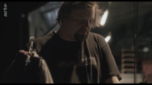 Un homme, possiblement un artiste ou un technicien, examine un vêtement sur un cintre lors des préparatifs d'une mise en scène ou d'une exposition. L'ambiance est celle d'un atelier ou d'une coulisse de théâtre.