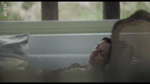 Une femme est allongée sur un canapé, les yeux fermés, dans une pièce baignée de lumière naturelle. L'ambiance est calme et contemplative.