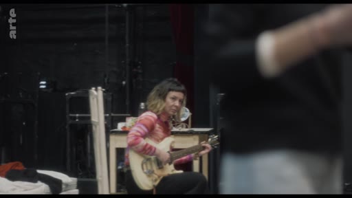 Une musicienne, vêtue d'un haut rayé coloré, joue de la guitare électrique dans une salle de répétition sombre. La scène capture un moment de travail artistique, probablement dans le cadre d'une préparation de spectacle ou d'un enregistrement.