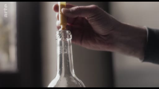 Une main manipule un tube de colle jaune pour l'insérer dans le goulot d'une bouteille en verre vide, illustrant une étape de préparation ou de bricolage.