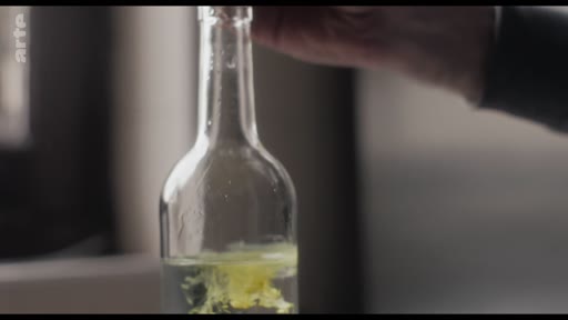 Une main verse un liquide jaune, probablement un ingrédient ou un colorant, dans une bouteille en verre transparente, illustrant une étape de préparation ou de mélange.