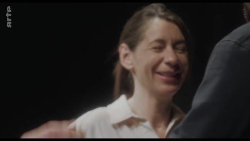Une femme souriante est filmée en gros plan dans une scène de vie quotidienne ou de performance. L'image capture une expression de joie ou de complicité lors d'un échange avec une autre personne.