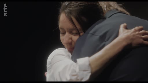Une femme en chemise blanche serre dans ses bras une personne vêtue d'un costume sombre, dans une scène intime et émotionnelle.