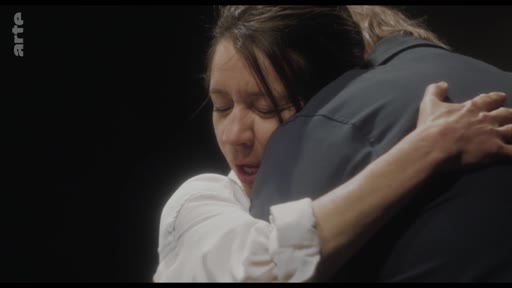 Une femme en chemise blanche serre une personne dans ses bras lors d'une scène dramatique ou théâtrale. L'image capture une émotion intense et une étreinte intime dans un contexte de performance.