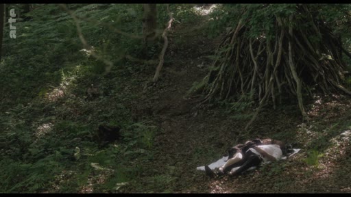 Deux individus sont allongés sur une couverture au sol dans une zone boisée, à proximité d'une structure faite de branches entrelacées. La scène se déroule en extérieur dans un environnement forestier.