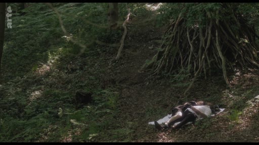 Un couple est allongé sur le sol dans une forêt, à proximité d'une structure en bois, dans une scène de fiction. L'image montre une ambiance naturelle et boisée.