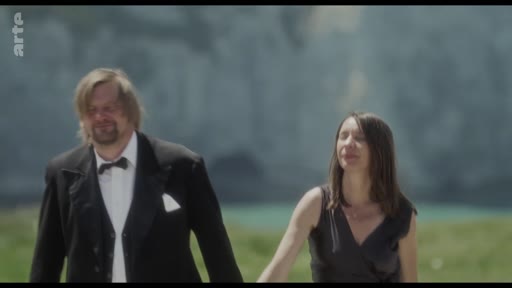 Un homme portant un costume formel avec un nœud papillon et une femme en robe noire marchent côte à côte dans un paysage naturel, probablement près d'une côte ou d'une plage.