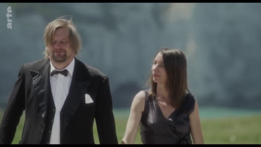 Un homme portant un costume formel avec un nœud papillon et une femme en robe de soirée marchent côte à côte dans un paysage extérieur, probablement lors d'un événement formel ou d'une cérémonie.