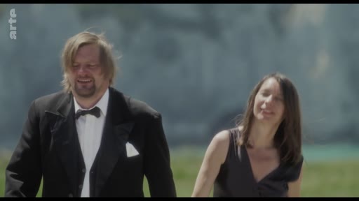 Un homme portant un costume avec un nœud papillon et une femme en robe noire marchent ensemble dans un paysage extérieur, probablement près d'une côte ou d'une plage.