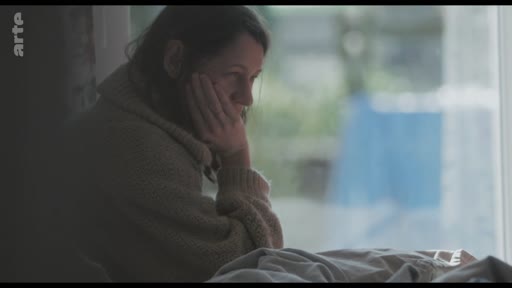 Une femme, le visage appuyé sur sa main, semble pensive ou préoccupée alors qu'elle regarde par la fenêtre dans une ambiance mélancolique.