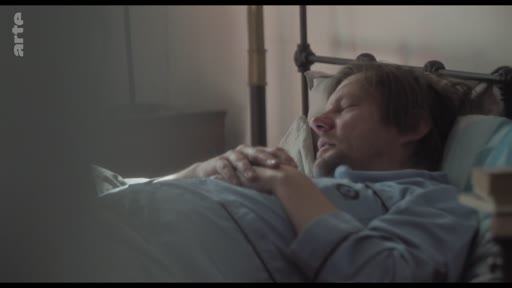 Un homme est allongé dans un lit, les mains jointes sur la poitrine, en train de dormir. La scène évoque un moment de repos ou de convalescence dans un cadre domestique.