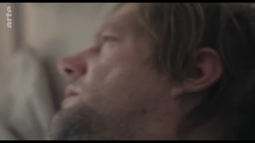 Un homme barbu, allongé et visiblement fatigué ou en proie à la douleur, est filmé en gros plan dans une scène évoquant la vulnérabilité ou la maladie.