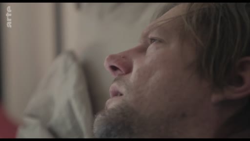 Un homme au visage marqué par la fatigue ou la maladie est filmé en gros plan alors qu'il est allongé, illustrant une situation de vulnérabilité ou de soin à domicile.