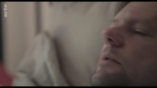 Un homme est filmé en gros plan alors qu'il dort, allongé dans un lit, dans une scène évoquant l'intimité ou le repos.