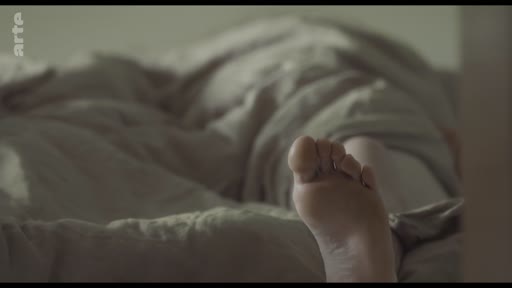 Un gros plan sur un pied dépassant d'une couette sur un lit, illustrant le thème de la solitude et de la vie domestique.