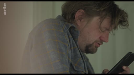 Un homme barbu, vêtu d'une chemise à carreaux, consulte son smartphone dans un moment de solitude. L'image illustre le thème de la communication isolée et de l'usage quotidien des technologies numériques.