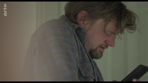 Un homme d'âge moyen, portant une chemise à carreaux, consulte son téléphone portable dans un moment d'isolement. L'image illustre le thème de la communication numérique et de la solitude.